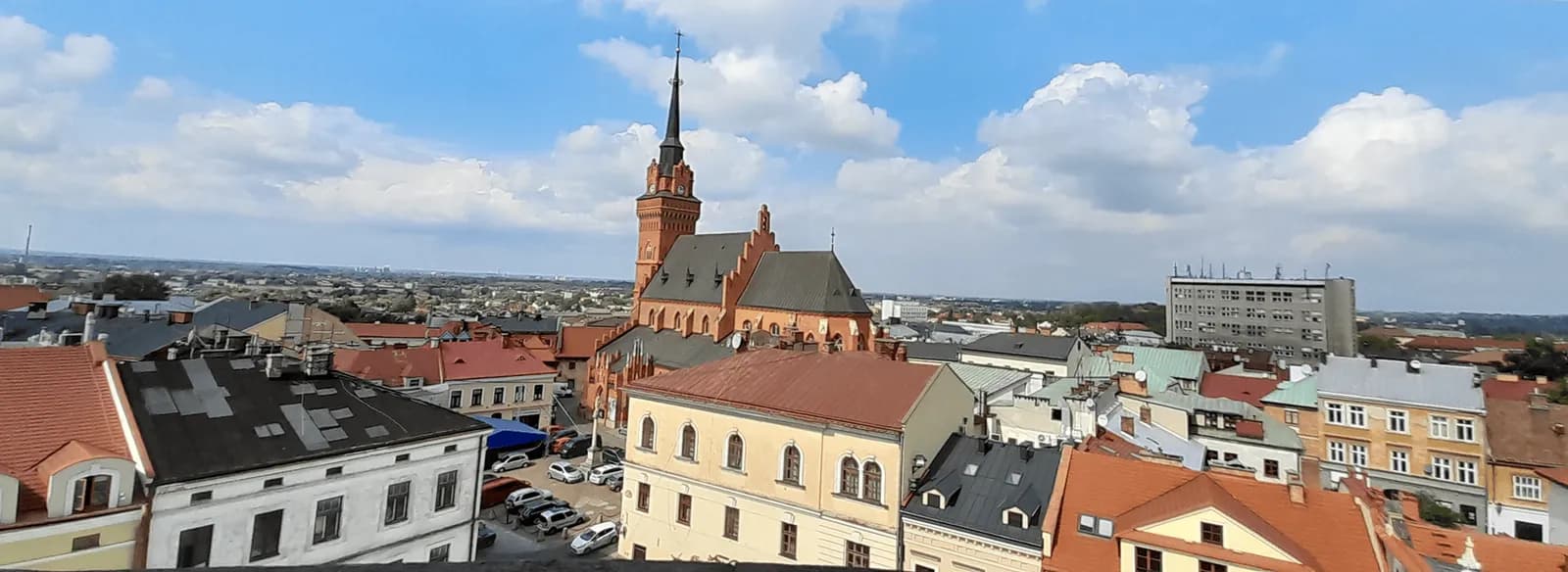 An aerial view of a European town with a prominent red-brick church spire dominating the skyline, surrounded by houses with red and dark roofs under a blue sky with white clouds.